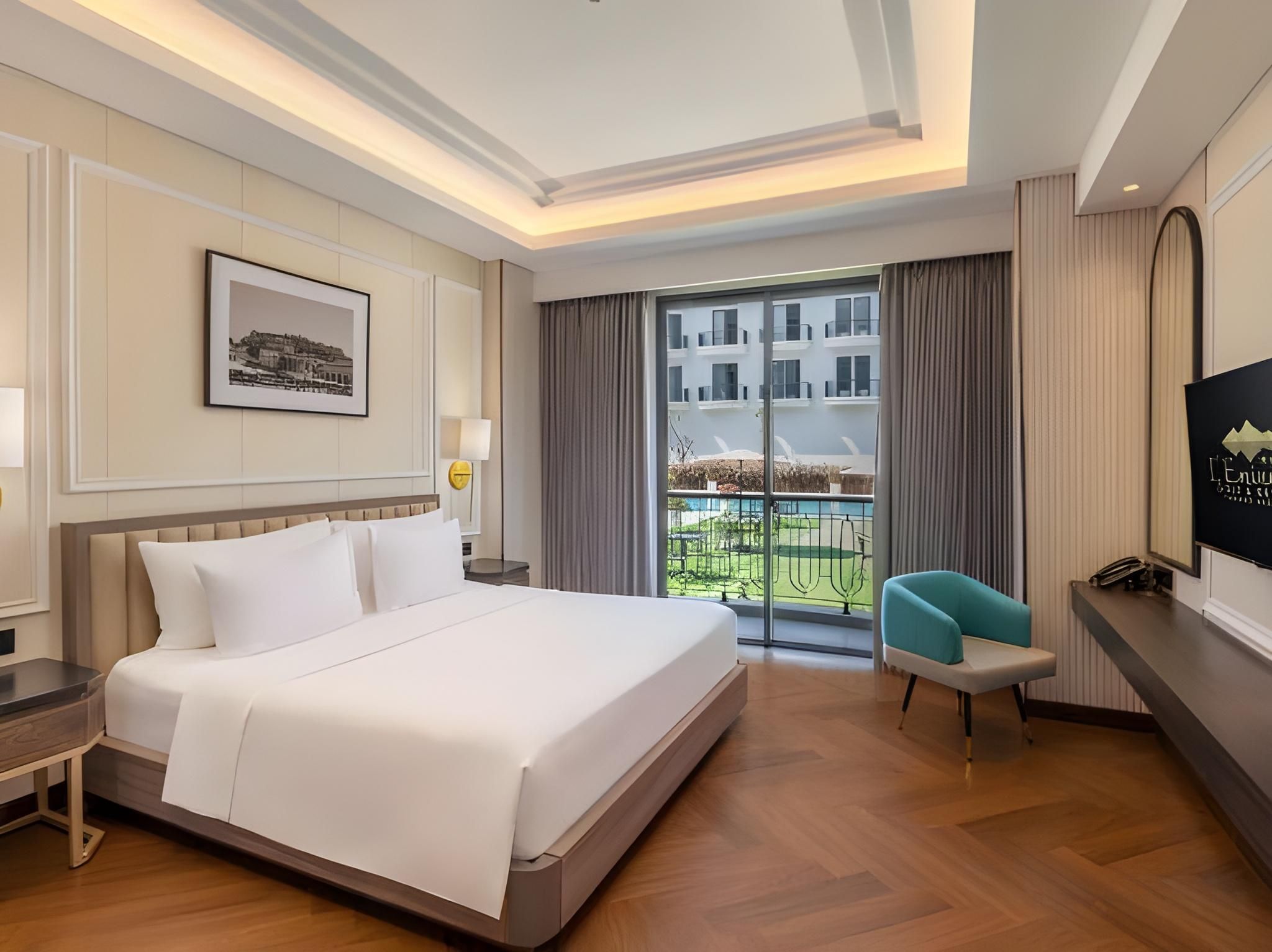 A hotel room with a large bed, a desk, a chair, a TV, and a balcony with a view of buildings.
