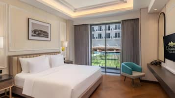 A hotel room with a large bed, a desk, a chair, a TV, and a balcony with a view of buildings.