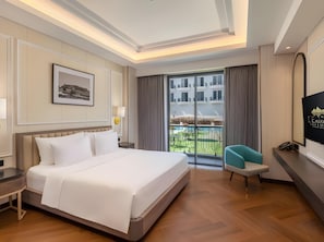 A hotel room with a large bed, a desk, a chair, a TV, and a balcony with a view of buildings.