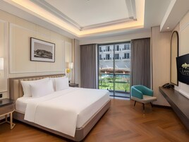 A hotel room with a large bed, a desk, a chair, a TV, and a balcony with a view of buildings.