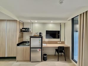 A compact kitchenette with a refrigerator, sink, and a microwave. There's a small table with a chair, a window with curtains, and a flat-screen TV mounted on the wall. - Rangsit Residence & Hotel (Thanyaburi)