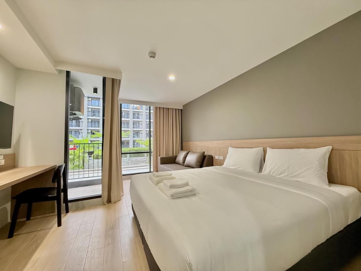 A hotel room with a bed, a desk, a chair, a TV, and a balcony with a view of buildings and greenery.