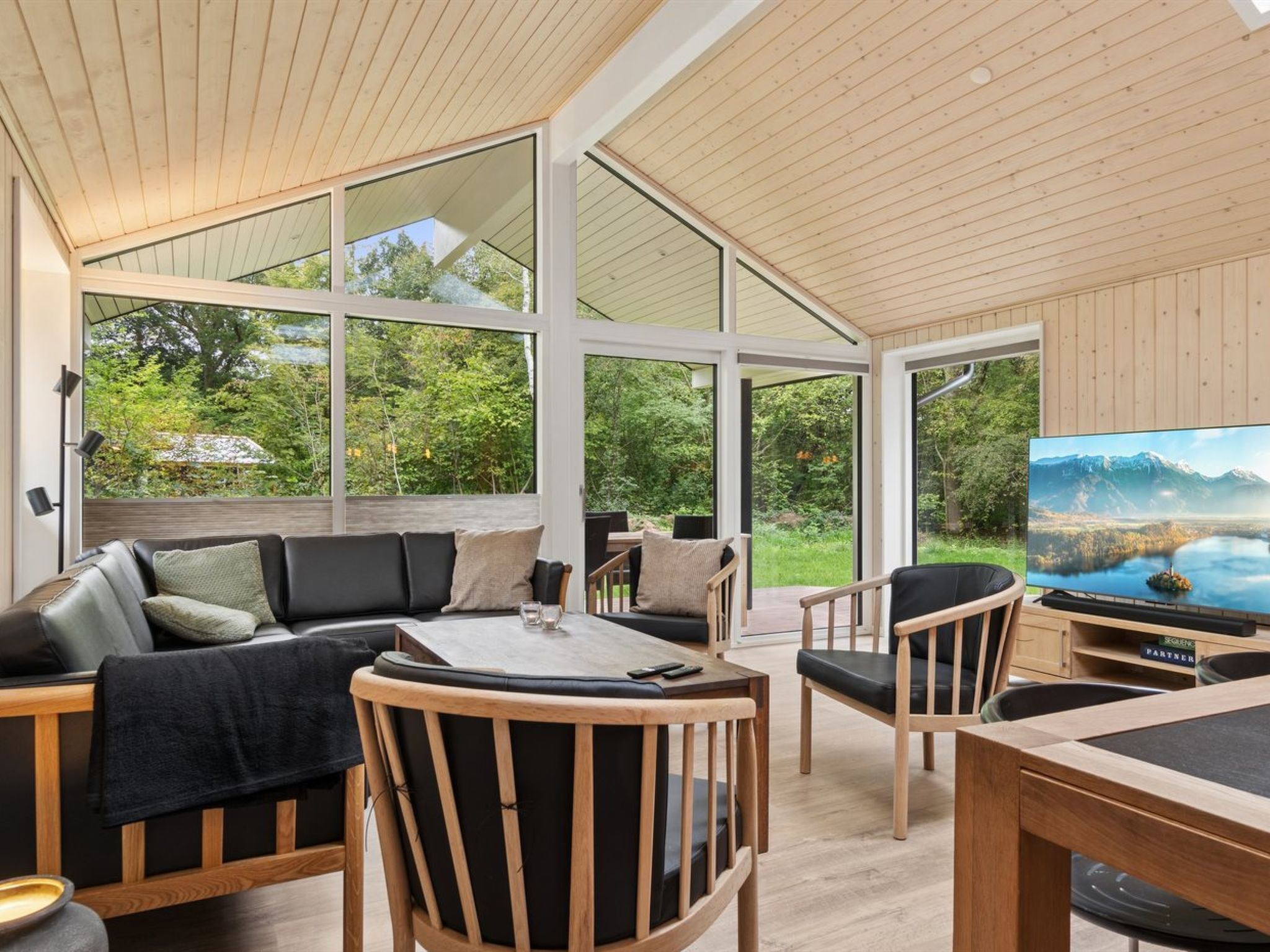 A modern living room with a large TV, wooden furniture, and large windows offering a view of the outdoors.