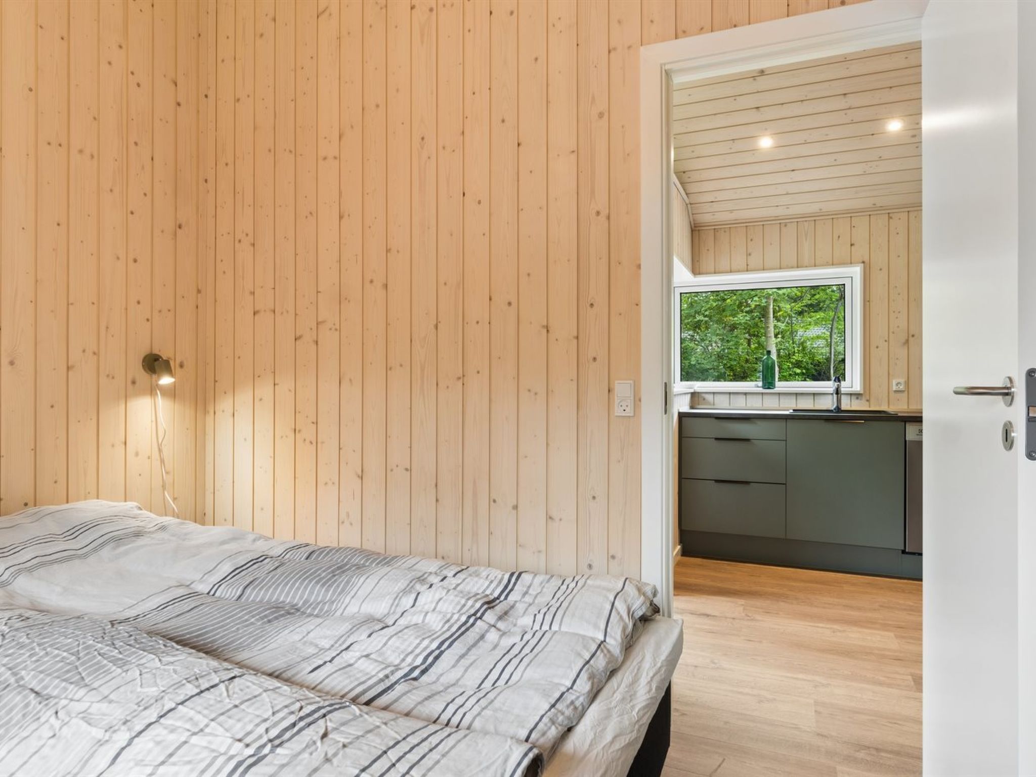 A bedroom with a bed, wooden walls, a window, and a view of a kitchen.