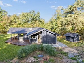 Ein modernes Haus mit Terrasse, Garage und einer kleinen Gartenhütte, umgeben von Bäumen und Rasen.