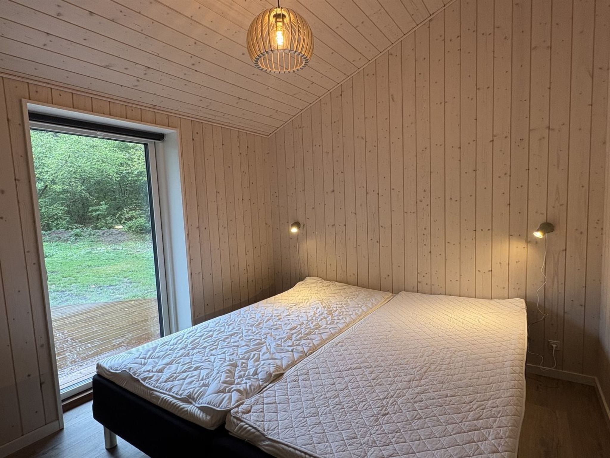 A bedroom with a large bed, wooden walls, a window, and a hanging light fixture.