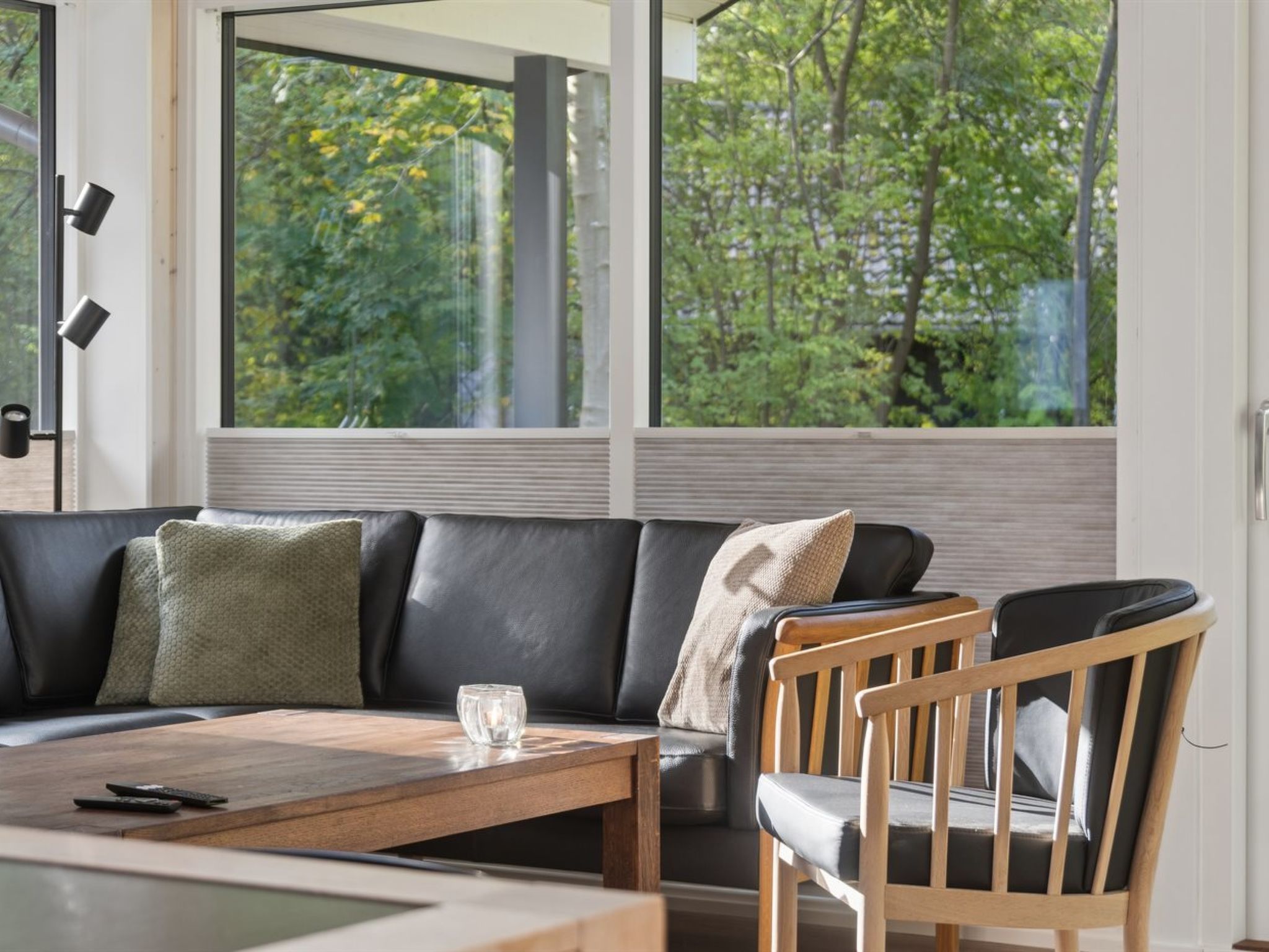 A modern living room with a leather sofa, a wooden coffee table, and a view of a forest through large windows.