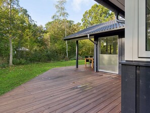 Une terrasse en bois mène à un bâtiment moderne doté de grandes fenêtres et d’un espace extérieur couvert.