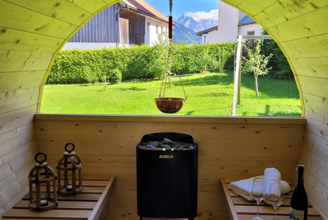 A wooden cabin with a view of a green lawn and mountains. Inside, there is a bed with a bottle of wine and glasses, a trash bin, and two lanterns.