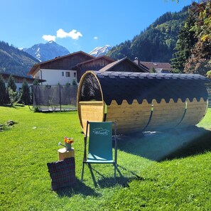 Un bote de madera con cabina, una silla y un florero sobre un campo cubierto de hierba, con casas y montañas al fondo.