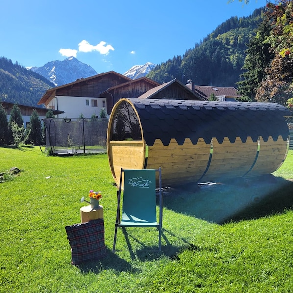A wooden boat with a cabin, a chair, and a flower vase on a grassy field with houses and mountains in the background.