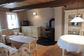 Une pièce chaleureuse avec un plafond en bois, une armoire blanche, un téléviseur, une table à manger recouverte d’une nappe blanche et une fenêtre avec des rideaux.