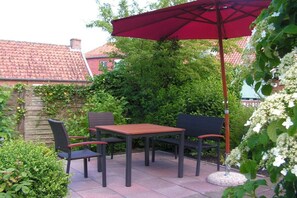 Eine Terrasse im Freien mit Tisch und Stühlen, einem roten Sonnenschirm und umgebender Bepflanzung.