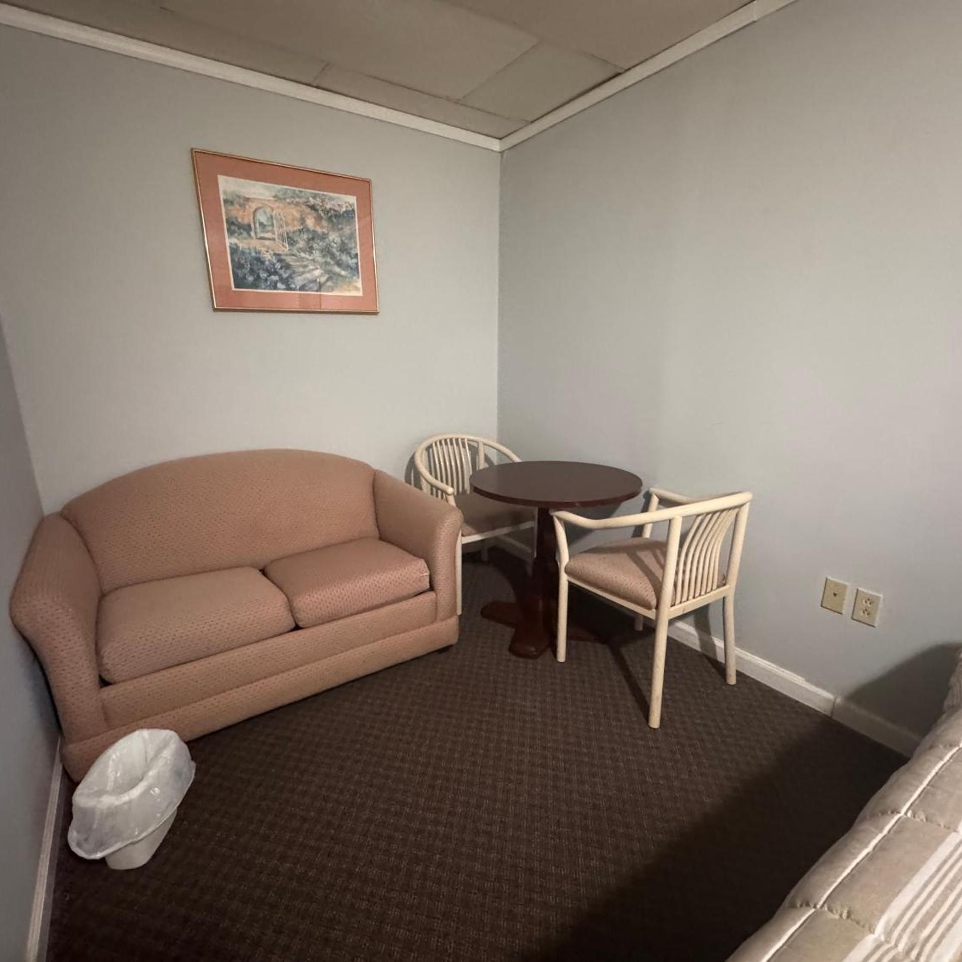 A room with a beige sofa, a round table, two white chairs, a framed picture on the wall, and a carpeted floor.