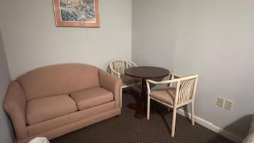 A room with a beige sofa, a round table, two white chairs, a framed picture on the wall, and a carpeted floor.