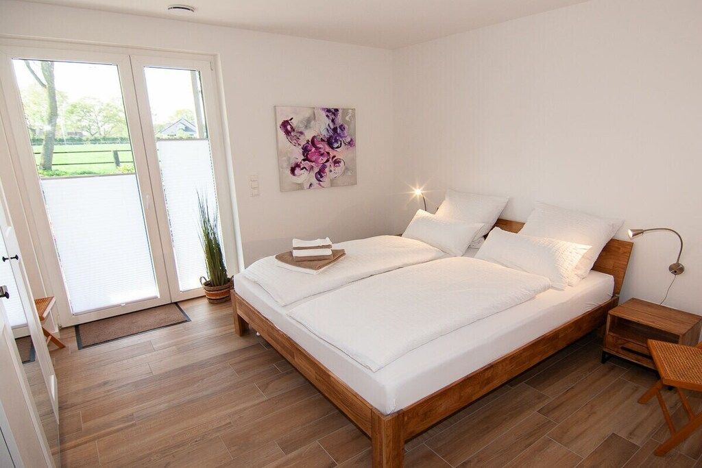 A bedroom with a wooden bed, white bedding, a bedside table, a nightstand, a wall-mounted lamp, and a door leading to an outdoor area.