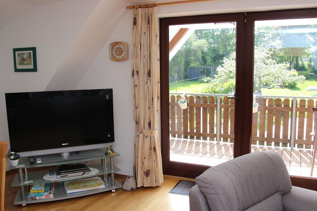 A living room with a television on a stand, a sofa, and a sliding glass door leading to a balcony with a wooden fence.