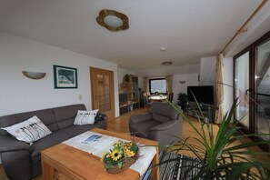 A living room with a sofa, armchairs, a coffee table with a floral arrangement, a television, and a bookshelf. - Holiday apartment with 1 bedroom (Cuxhaven)