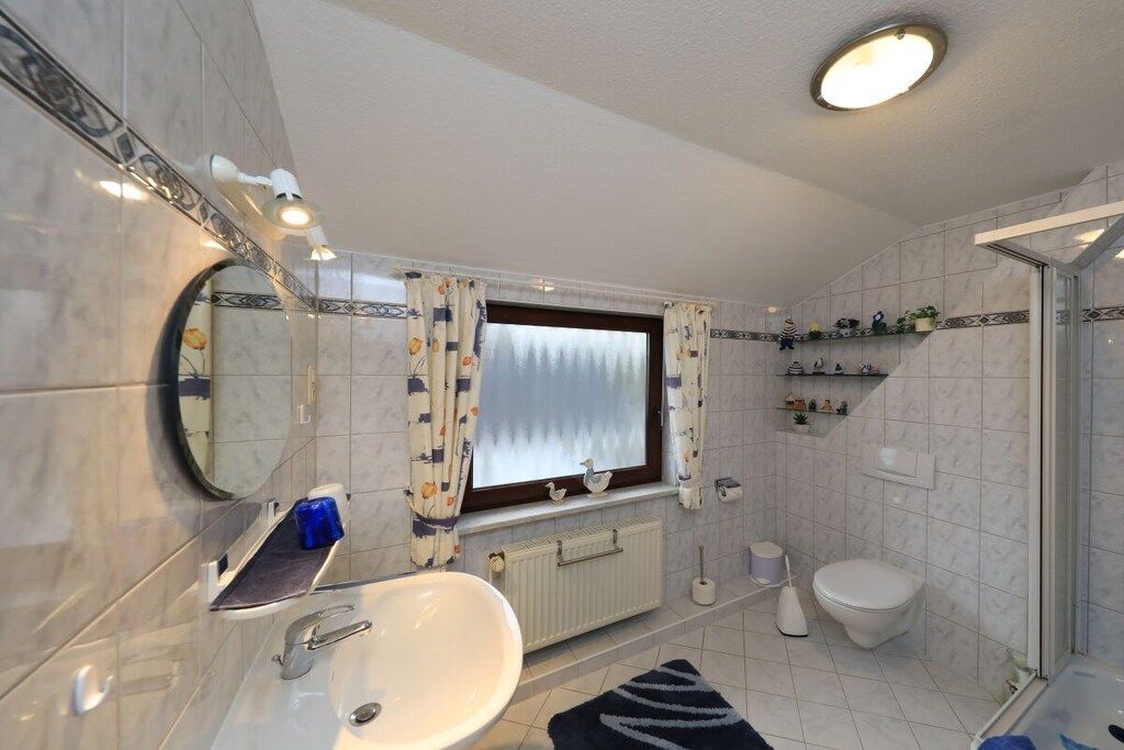 Ein Badezimmer mit weißem Waschbecken, einem Spiegel, einem Fenster mit Vorhängen, einer Duschkabine und einer Toilette.