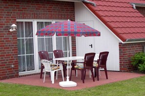 A patio with a table and chairs, a plaid umbrella, and a brick building with a white door. - Holiday home family Leitner (Burhave)
