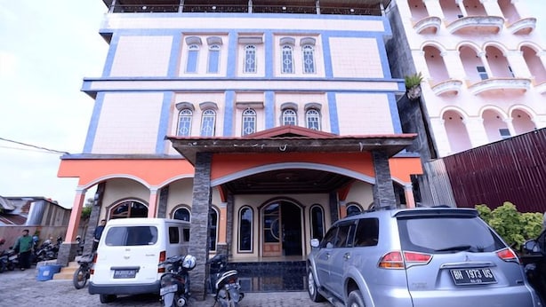 Un edificio de varios pisos con una entrada destacada, vehículos estacionados y otra estructura contigua con balcones.
