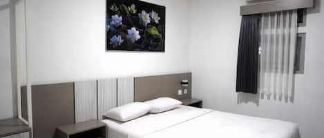 A hotel room with a bed, a nightstand, a painting on the wall, and an air conditioning unit.