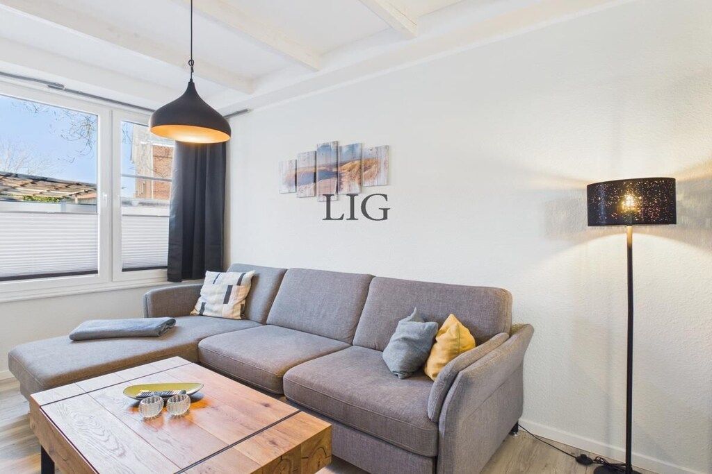 Ett modernt vardagsrum med en grå soffa, ett soffbord i trä, en svart hänglampa, en golvlampa och konstverk på väggen.