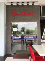 Entrada do Hotel Viva, com porta de vidro, tapete de boas-vindas e uma cadeira vermelha do lado de fora.