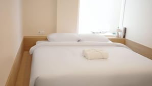 Uma cama de solteiro com roupa de cama branca e uma toalha bem dobrada, uma mesinha de cabeceira de madeira e uma janela que traz luz natural.