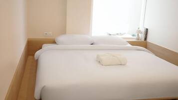A single bed with white bedding and a neatly folded towel, a wooden bedside table, and a window providing natural light.