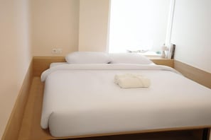 Ein Einzelbett mit weißer Bettwäsche und einem ordentlich gefalteten Handtuch, ein Nachttisch aus Holz und ein Fenster, das für natürliches Licht sorgt.