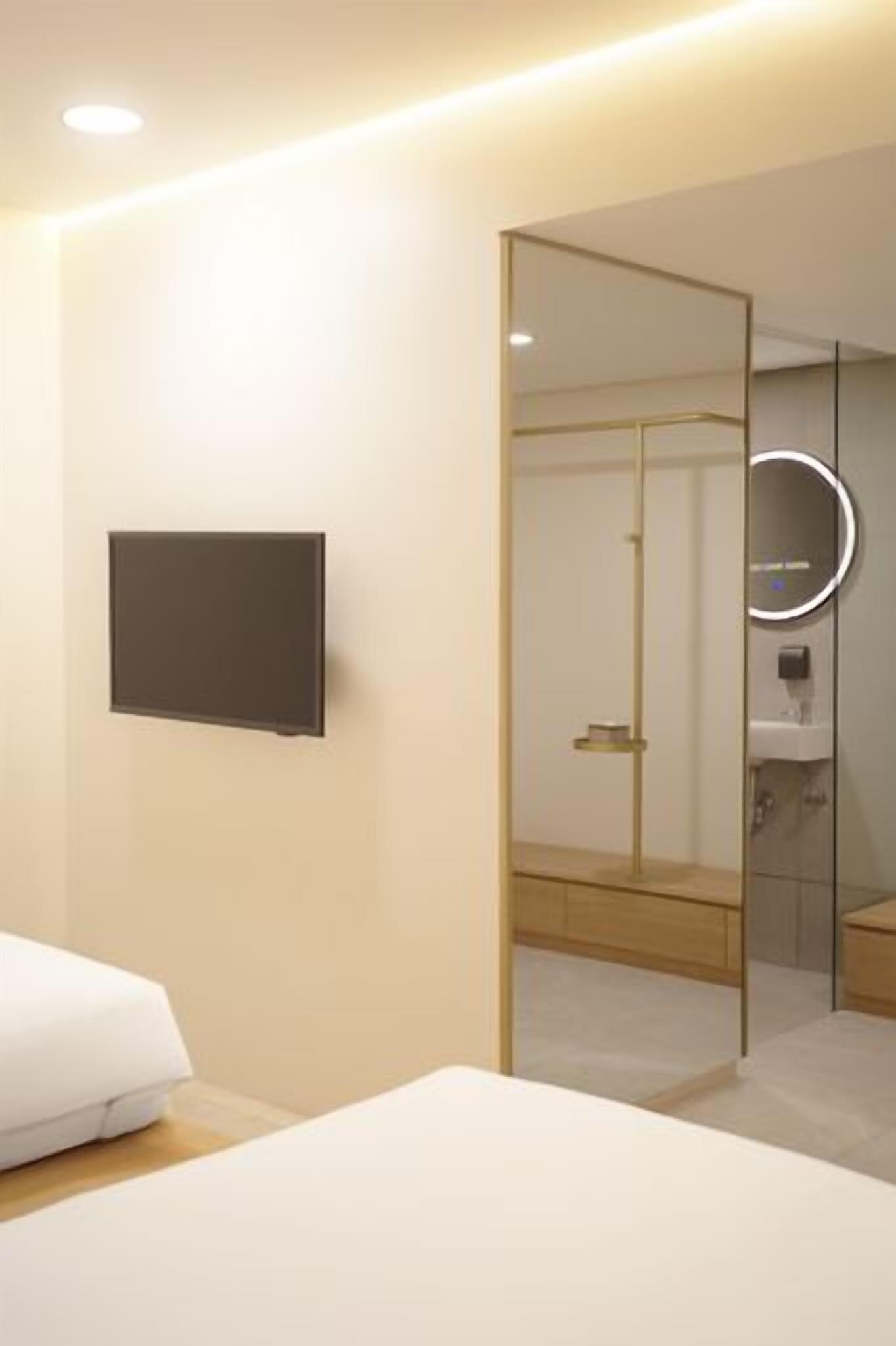 Une chambre d’hôtel moderne équipée d’un lit, d’un téléviseur à écran plat, d’un miroir et d’une salle de bain visible à travers une paroi en verre.