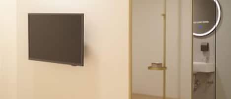 Une chambre d’hôtel moderne équipée d’un lit, d’un téléviseur à écran plat, d’un miroir et d’une salle de bain visible à travers une paroi en verre.