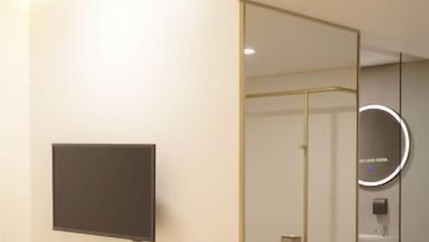 Quarto de hotel moderno com cama, televisão de tela plana, espelho e banheiro visível por meio de um painel de vidro.