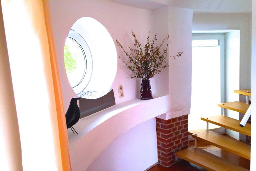 Una habitació amb una paret blanca corbada, una finestra circular, una llar de foc de maó i una escala de fusta.