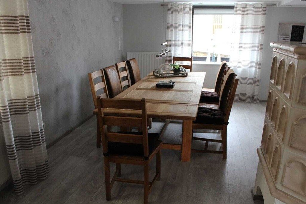 Un coin repas avec une table et des chaises en bois, un radiateur et une vue sur l’extérieur à travers une fenêtre ornée de rideaux.
