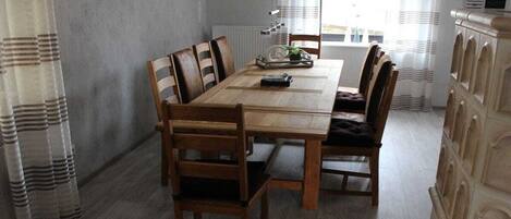 Un coin repas avec une table et des chaises en bois, un radiateur et une vue sur l’extérieur à travers une fenêtre ornée de rideaux.