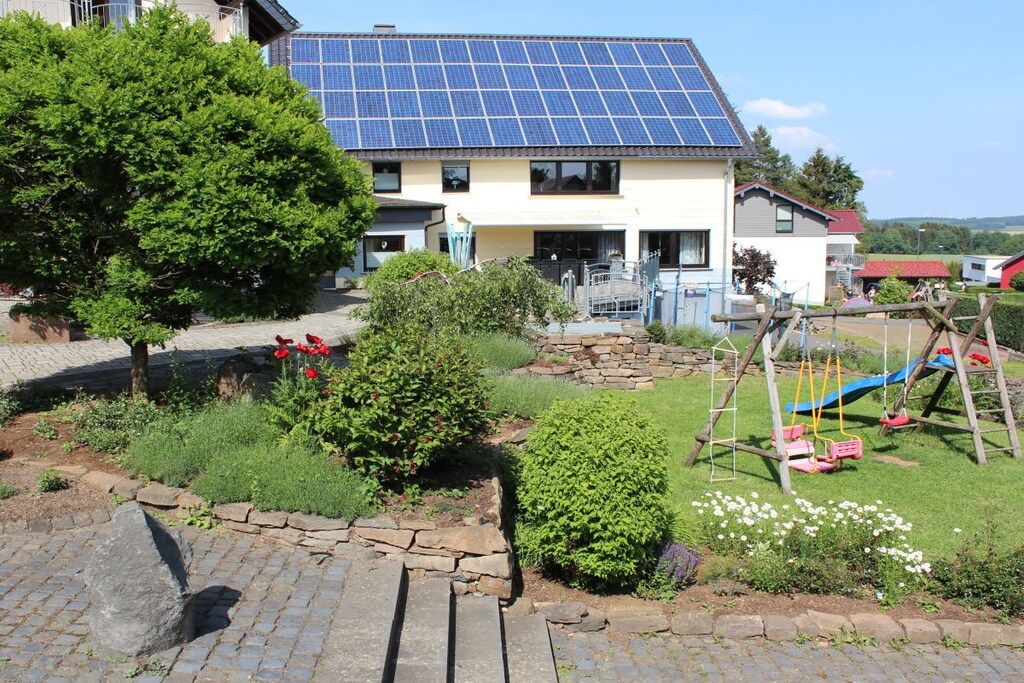 A residential building with a solar panel roof, a garden with a swing set, and a paved driveway.