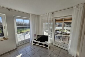 A modern living room with a flat-screen TV on a white stand, large windows with white curtains, and a view of a patio area. - Holiday apartment with 2 bedrooms (Wenkendorf)