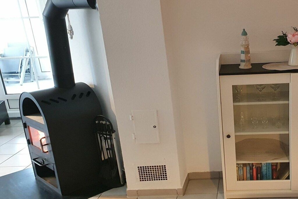 Quarto com fogão a lenha preto, armário branco com portas de vidro e uma pequena mesa com um vaso e livros.