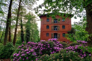 A red brick building with multiple windows and shutters, surrounded by lush greenery and purple flowers. - Time out The holiday home (Müden\/Örtze)