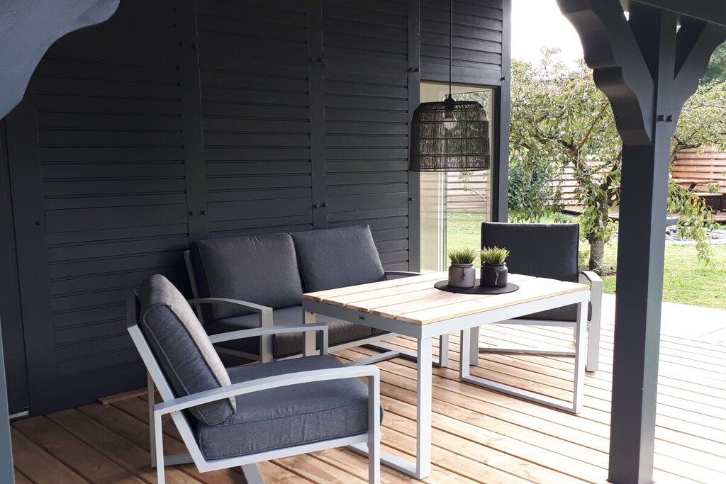 An outdoor patio with a wooden table and two chairs, a hanging light fixture, and a view of the backyard.