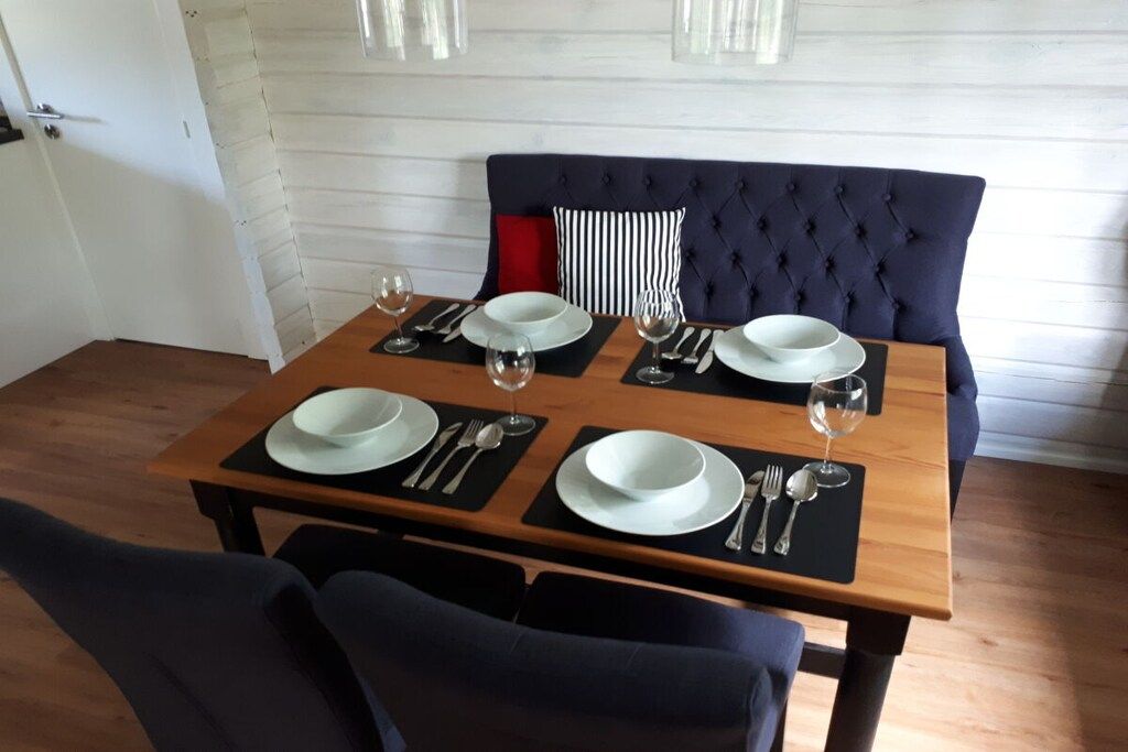 A dining table set with four plates, cutlery, and wine glasses, positioned in front of a dark upholstered bench with a striped pillow.