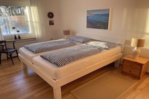 Ein Doppelbett mit gestreifter Bettwäsche, ein Nachttisch mit Lampe, ein kleiner Holznachtschränkchen, ein Stuhl und ein Fenster mit Vorhängen.