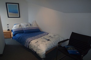Ein Einzelbett mit gestreifter Bettdecke und Kissen, ein Nachttisch mit Lampe, ein gerahmtes Bild an der Wand und ein Sessel mit einem Buch.