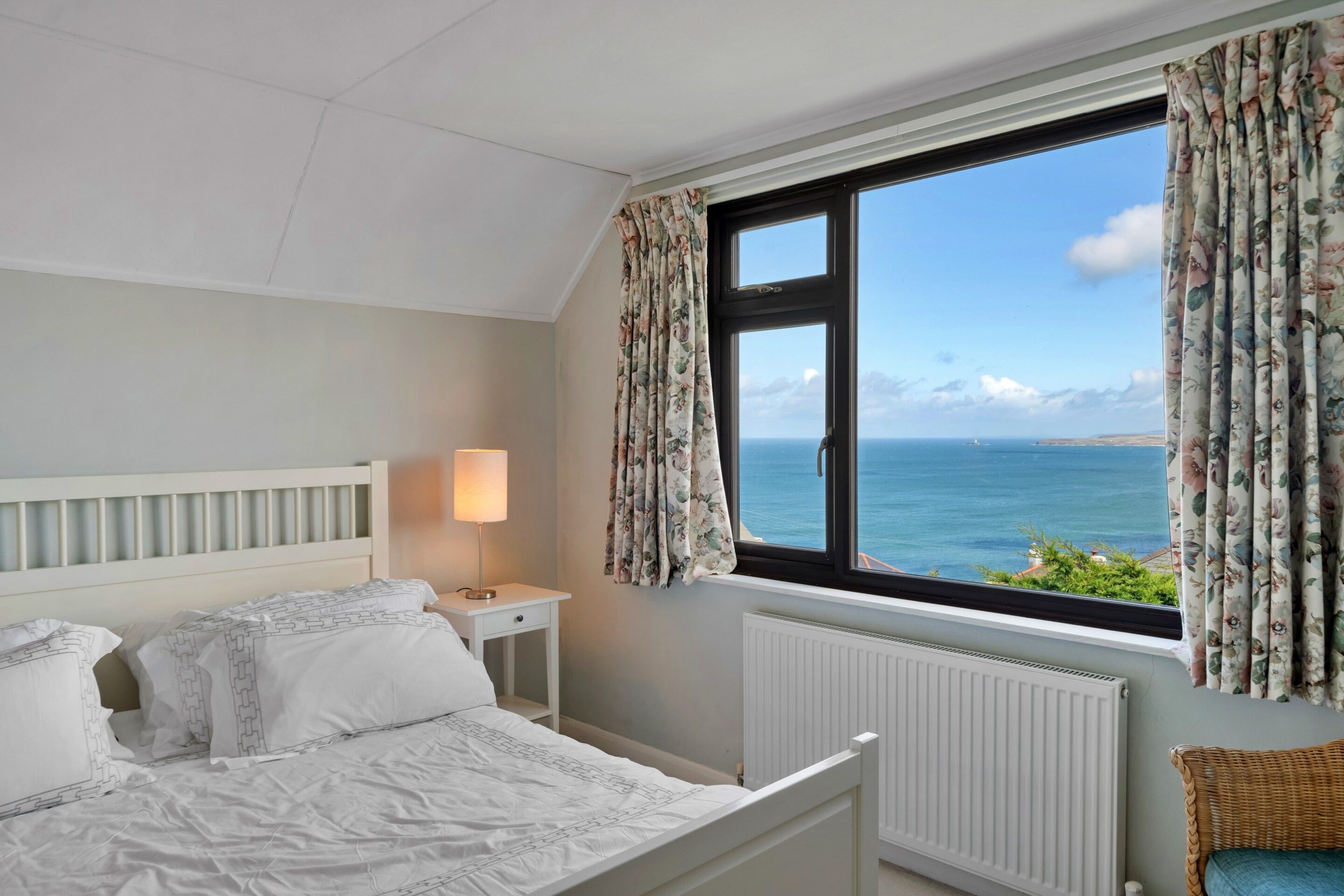 A bedroom with a bed, a nightstand, a lamp, a window with curtains, and a view of the sea.