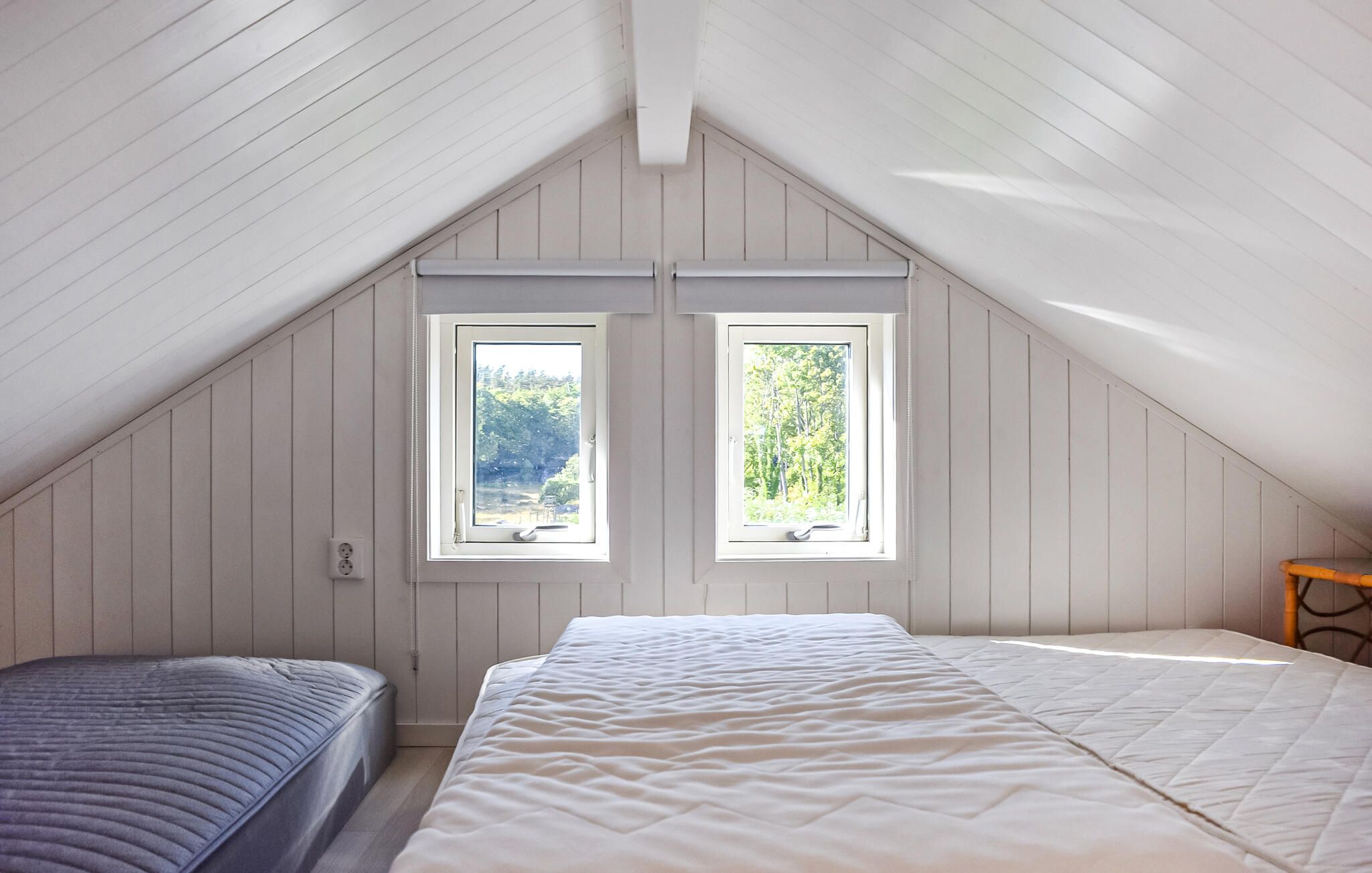 Ein Schlafzimmer mit geneigter Decke, zwei Fenstern, einem Bett und einem Sessel.