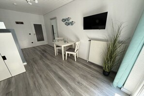 A room with a dining table, chairs, a potted plant, a radiator, and a television mounted on the wall. - Friesenkoje Horumersiel - bunk 3 (Wangerland)