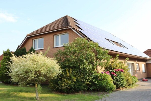 Een huis met een schuin dak, zonnepanelen, en een keurig onderhouden tuin met verschillende struiken en een kleine boom.