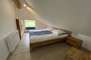A bedroom with a sloped ceiling, a wooden bed, a nightstand, and a window with a view of greenery. - Holiday apartment with 2 bedrooms (Pilsum)